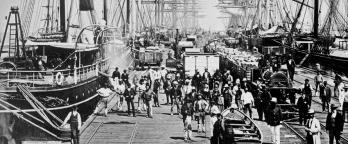 Black and white photo of people on pier with tall ships and pier donkey.